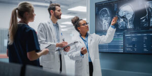 Reunión de laboratorio en un hospital de ciencias médicas: un equipo diverso de neurólogos, neurocientíficos y neurocirujanos consultan una pantalla de televisión que muestra una resonancia magnética con imágenes cerebrales y hablan sobre métodos de tratamiento y nuevos fármacos para la cura.