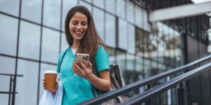 Una alegre profesional de la salud, vestida con un uniforme verde azulado, disfruta de un momento afuera de un centro médico, con un café en la mano, mientras navega en su teléfono inteligente.