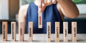 Mano de empresario colocando o tirando fichas de dominó de madera con el texto MARCA y Marketing, Publicidad, Logotipo, Diseño, Estrategia, Identidad, Confianza y Valores. Concepto de desarrollo de producto.