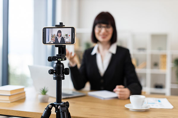 Mujer caucásica con traje negro grabando un videoblog en una oficina usando un teléfono inteligente en un trípode.