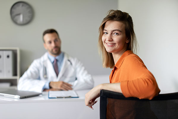 Retrato de una paciente joven feliz que se siente satisfecha con el servicio de buena calidad en la clínica, sentada en una consulta con un médico en el hospital y sonriendo a la cámara.