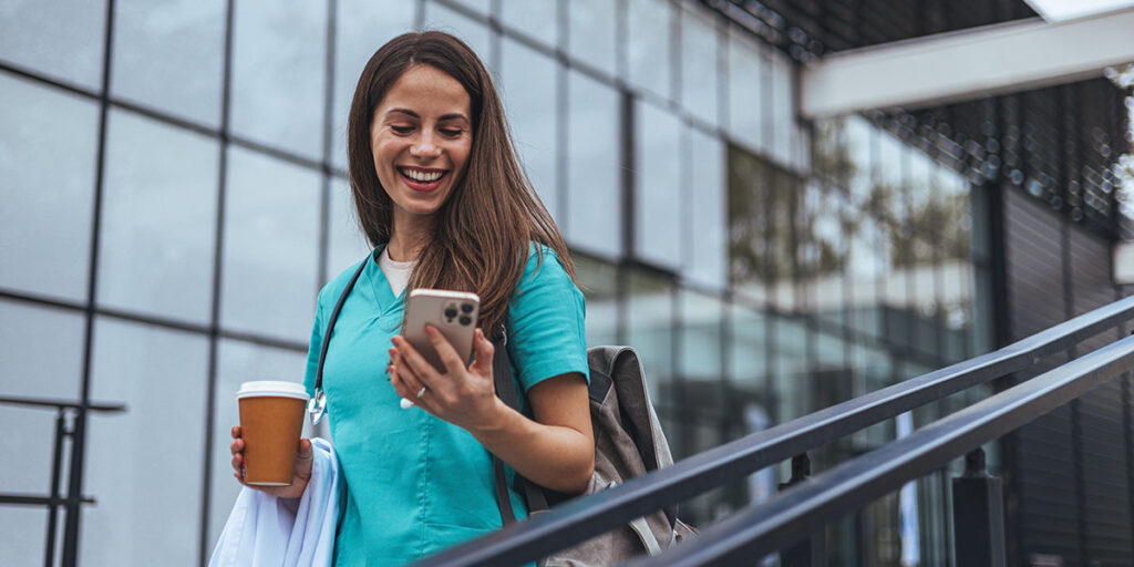 Una alegre profesional de la salud, vestida con un uniforme verde azulado, disfruta de un momento afuera de un centro médico, con un café en la mano, mientras navega en su teléfono inteligente.