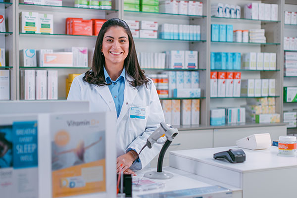 Mostrador de caja de farmacia: Retrato de una hermosa joven farmacéutica latina que mira a la cámara y sonríe encantadoramente. Tienda con medicamentos, fármacos, vitaminas y productos para el cuidado de la salud.
