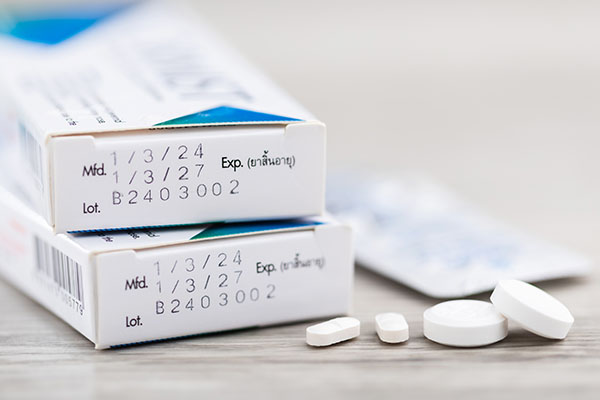 Primer plano del código de producción y la fecha de caducidad impresos en el lateral de una caja de medicamentos, con pastillas colocadas sobre una mesa. La imagen resalta la importancia de comprobar los medicamentos antes de consumirlos.