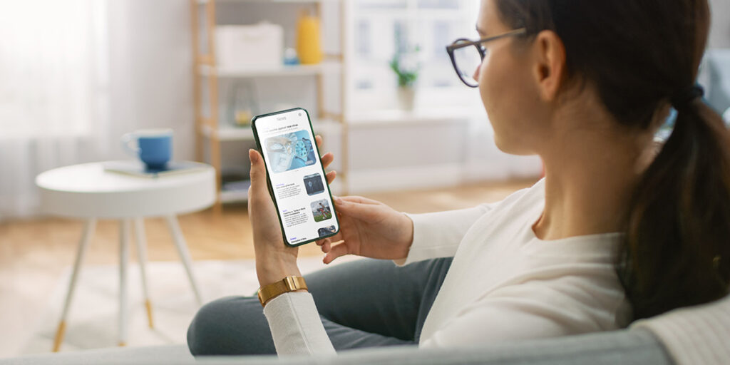 Una joven en su casa usa su teléfono inteligente para navegar y leer noticias sobre avances tecnológicos. Está sentada en un sofá en su acogedora sala de estar. Toma desde atrás.